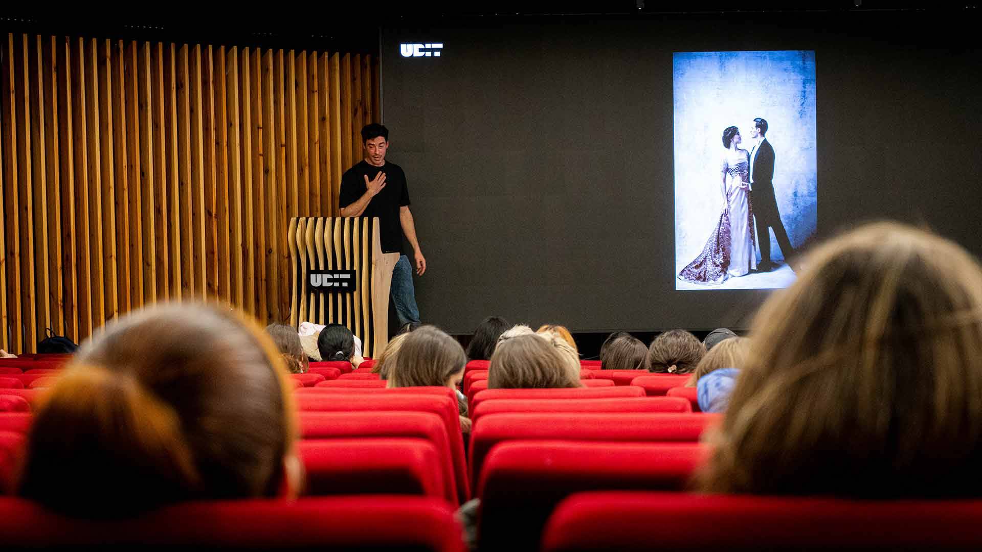 Un presentador habla frente a un público mientras muestra una imagen en una pantalla grande.