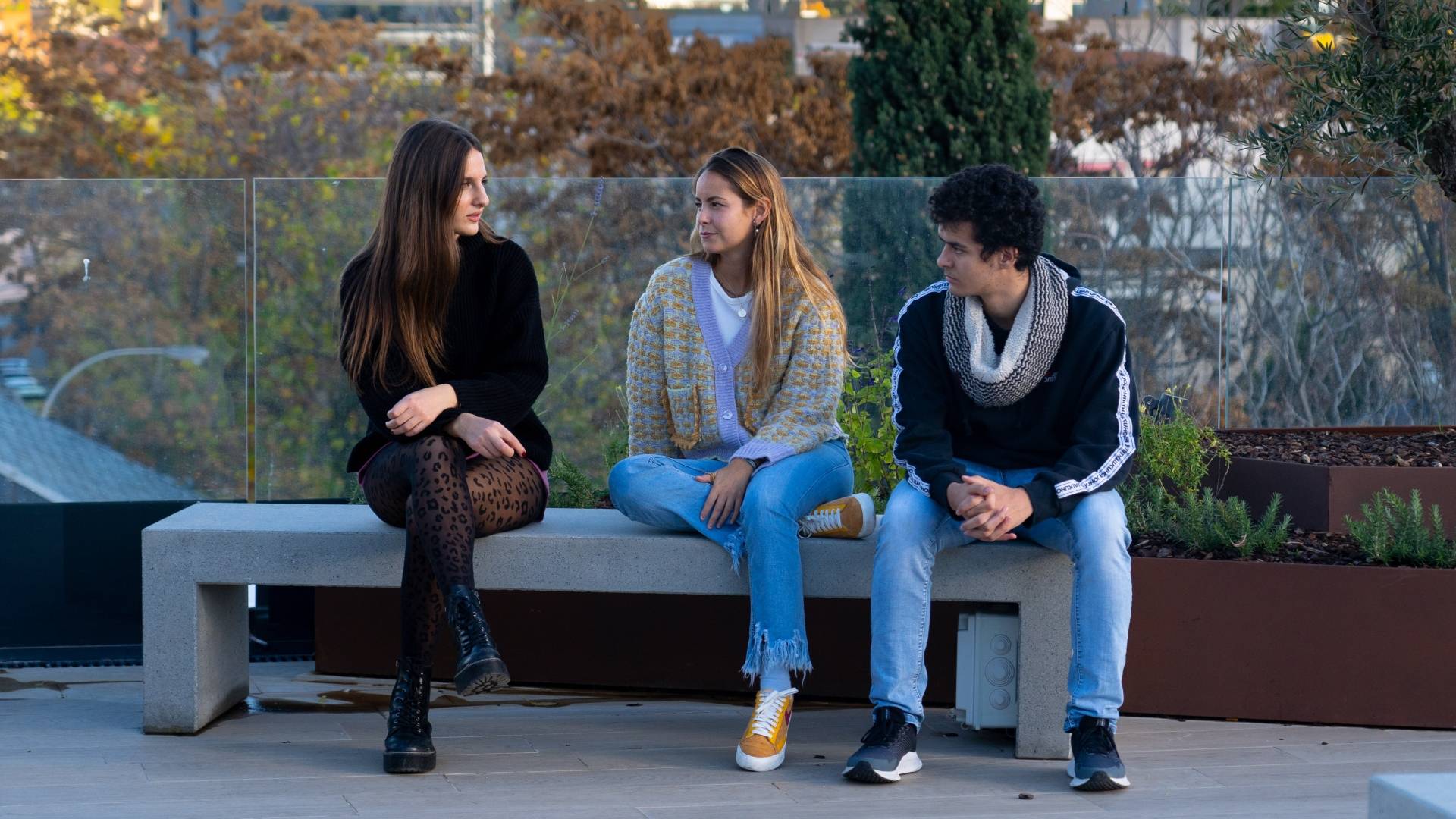 Tres jóvenes sentados en una banca al aire libre conversando entre ellos.
