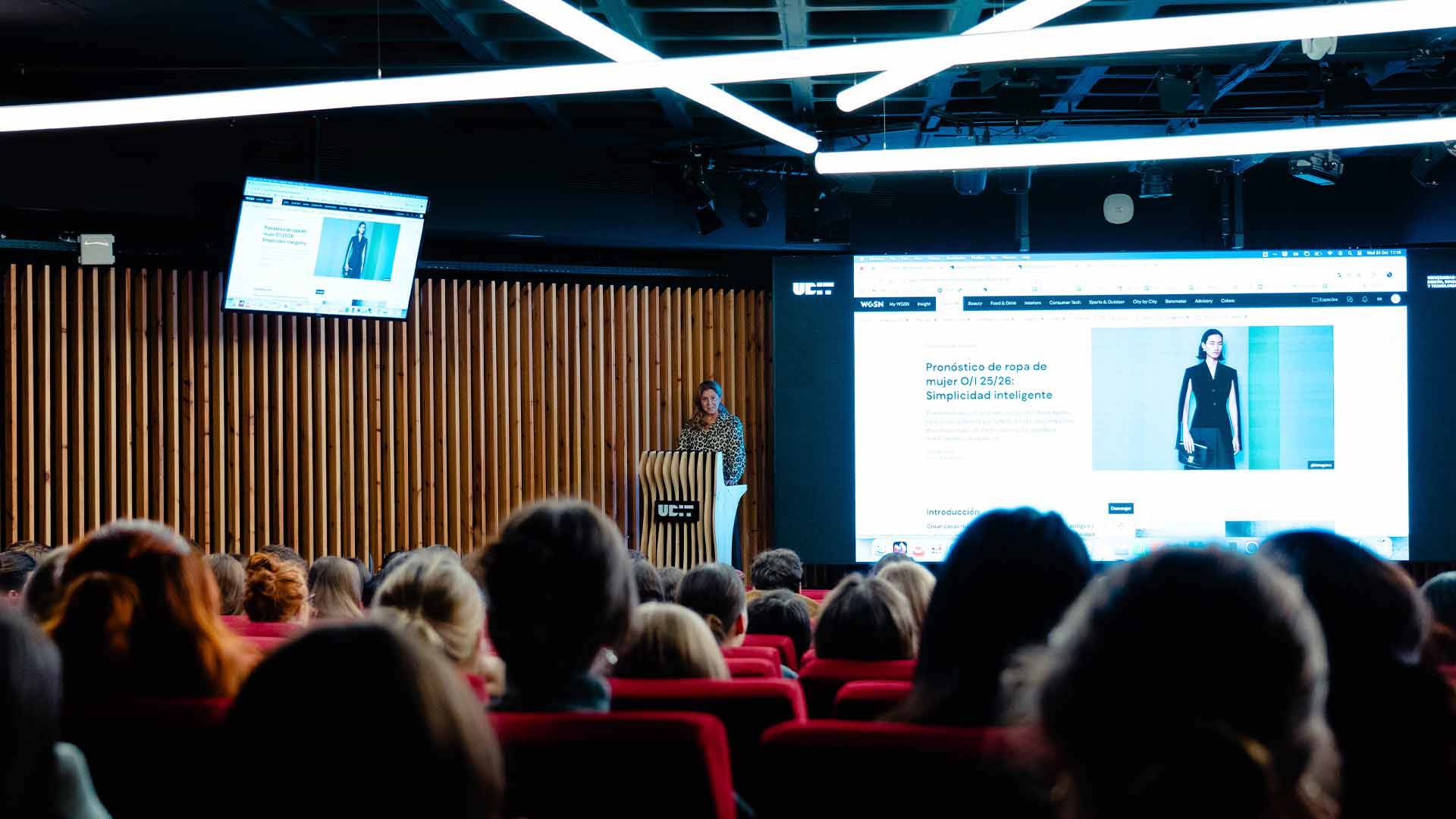 Una mujer presenta en un auditorio mientras la audiencia escucha atentamente.