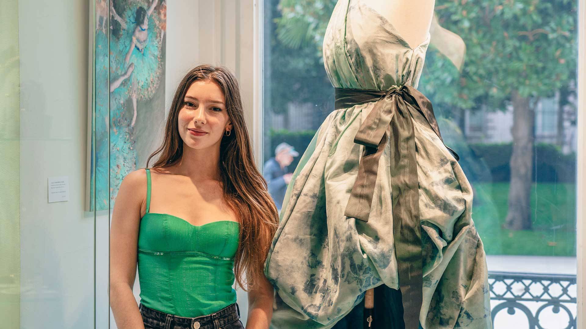 Una joven posa junto a un maniquí que lleva un vestido elegante en una galería.