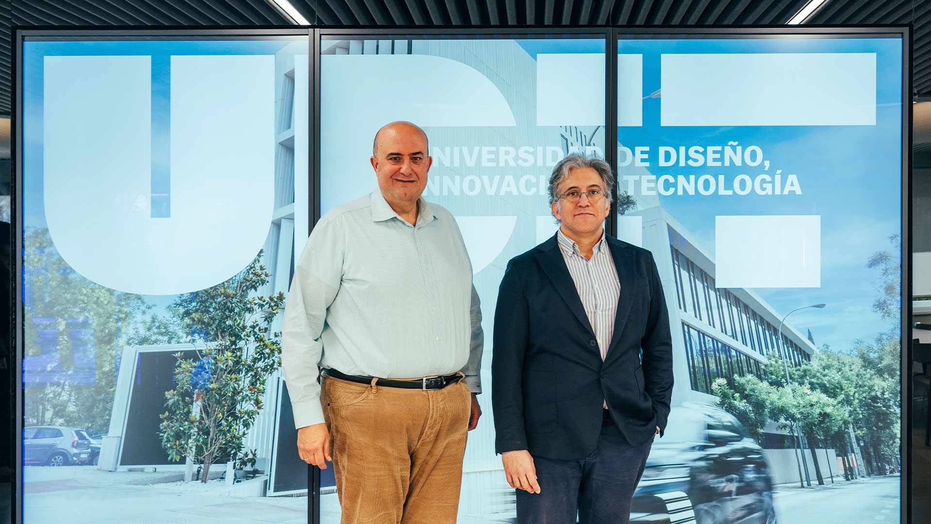 Dos hombres posan frente a un fondo con el logo de una universidad de diseño y tecnología.