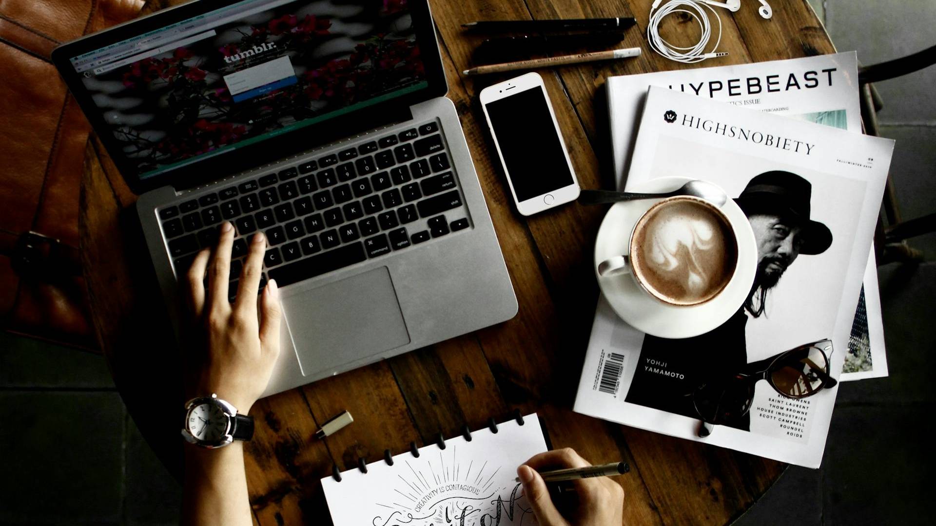 Una persona trabaja en una computadora portátil mientras disfruta de un café en una mesa con artículos de diseño y tecnología.