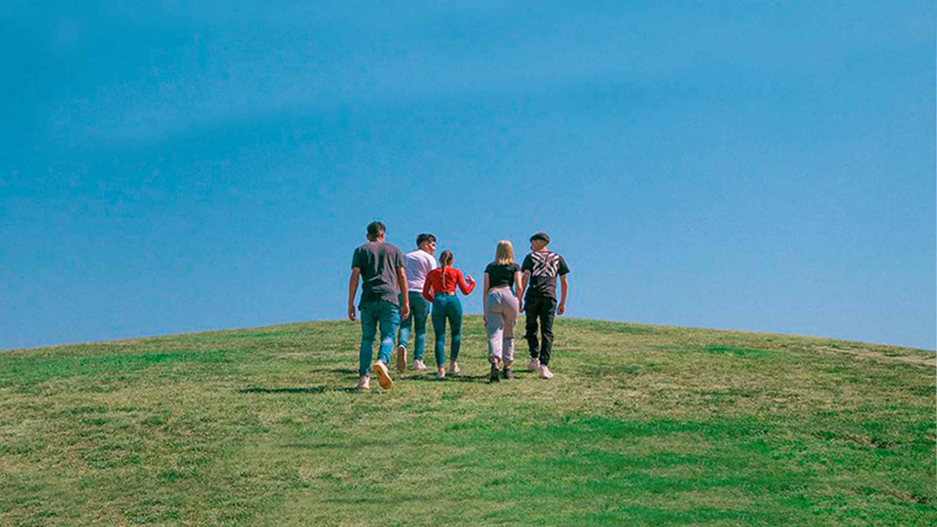 Un grupo de cinco personas camina por una colina verde bajo un cielo azul.