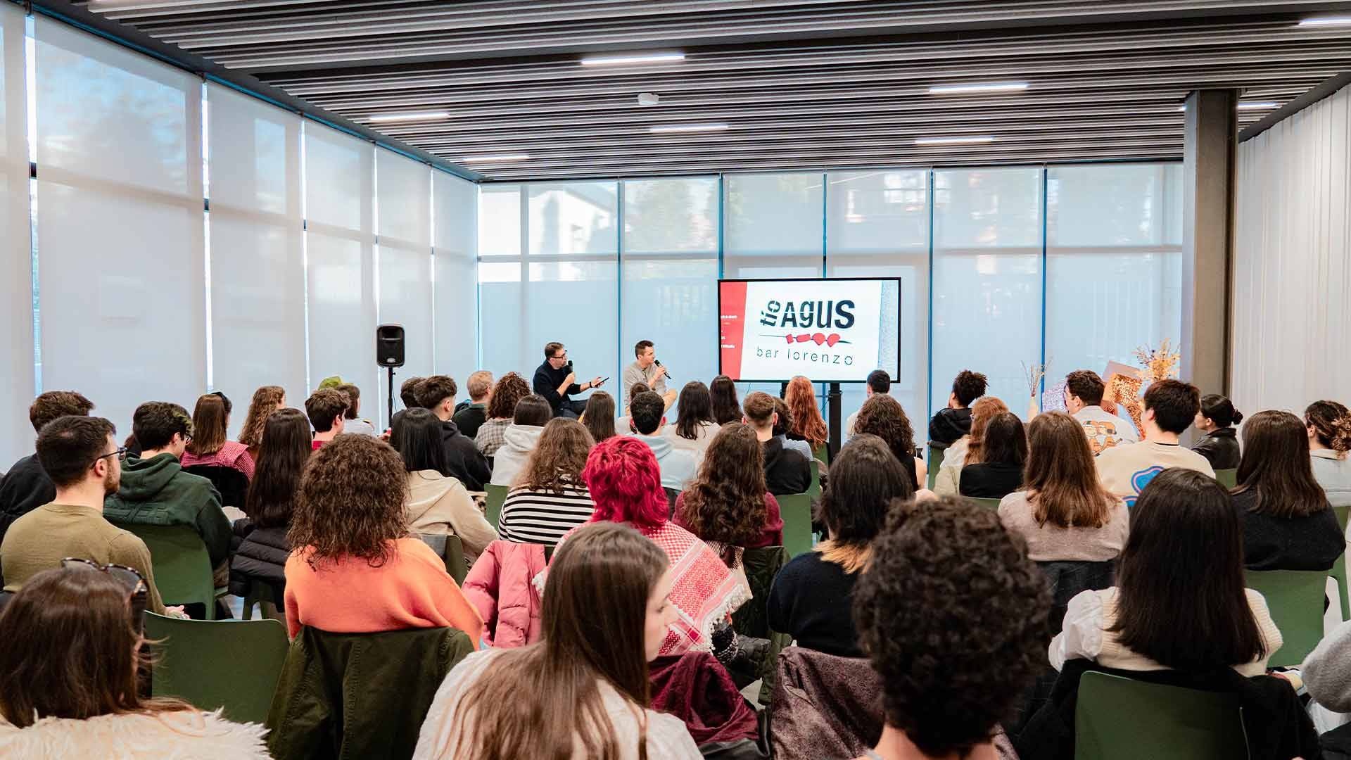 Un grupo de personas asiste a un evento o conferencia en una sala con pantallas y un presentador en el escenario.