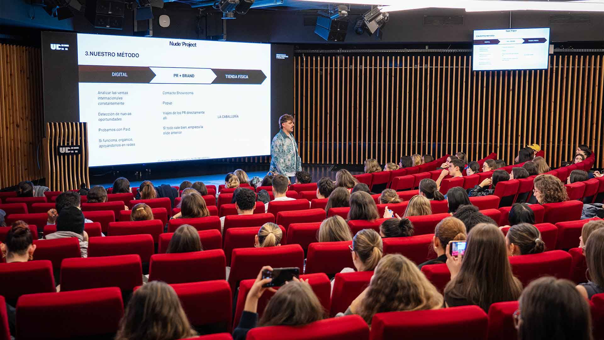 Una presentación en un auditorio con un ponente y un público sentado en butacas rojas.