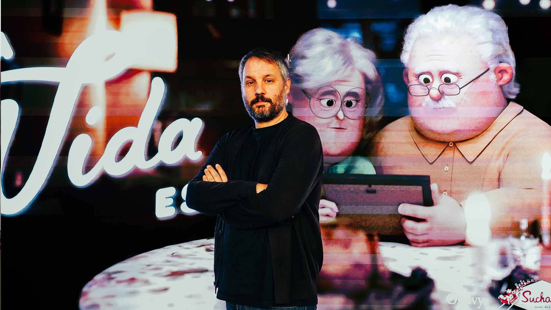 Un hombre posando frente a un fondo animado que muestra a dos personajes de estilo caricaturesco y el texto 'Vida'.