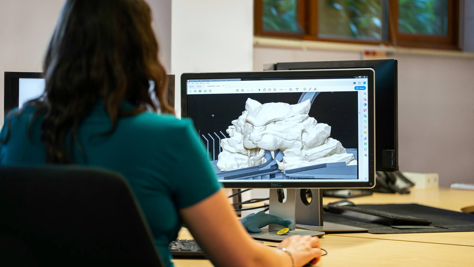 Una persona sentada frente a un ordenador que muestra un modelo tridimensional en la pantalla.