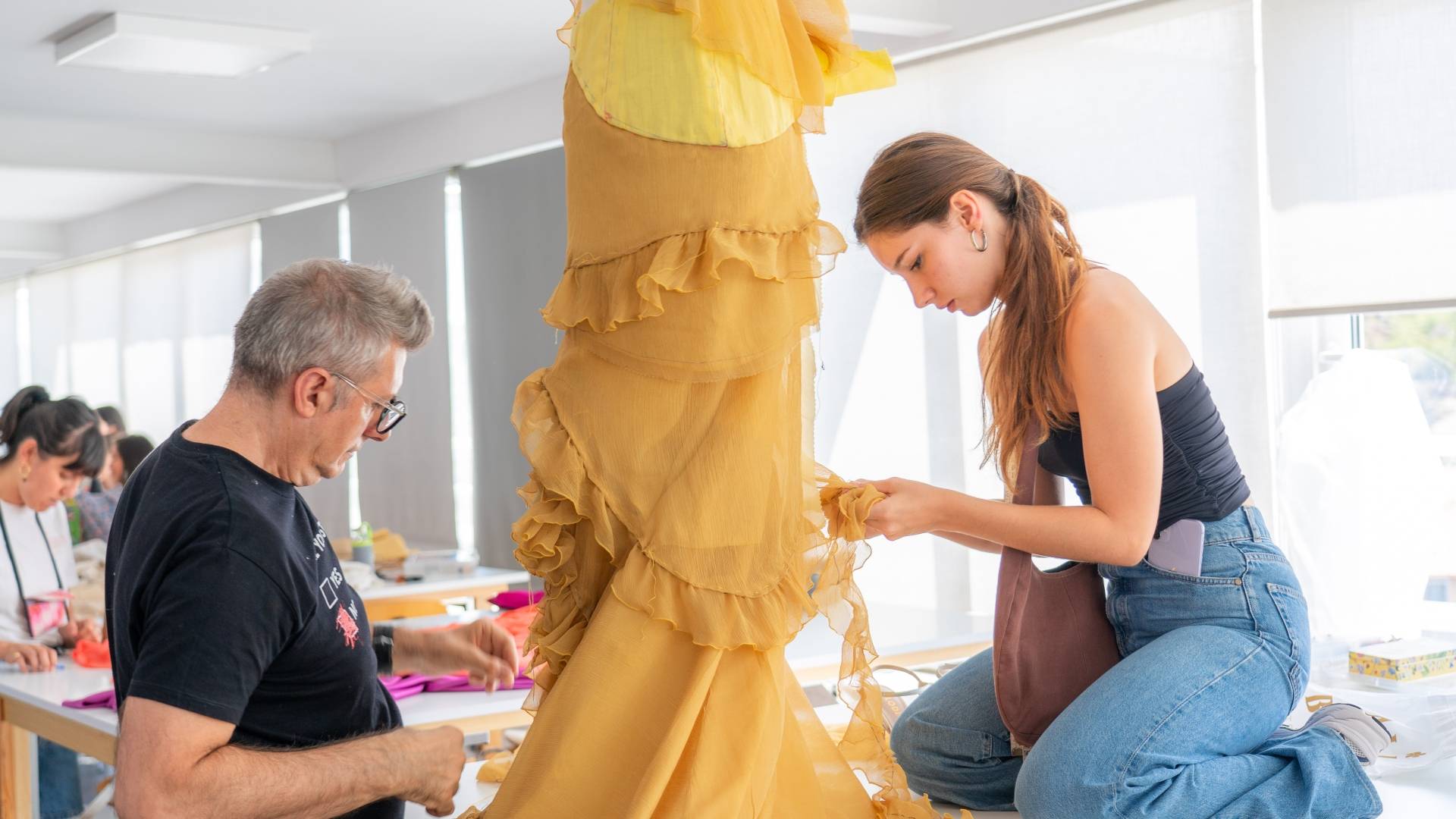 Una mujer trabaja en un vestido de tela amarilla mientras un hombre la observa en un taller de costura.