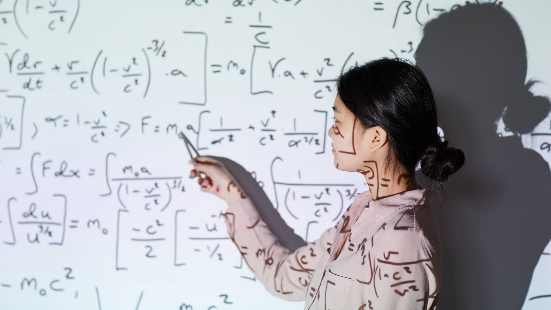 Una mujer señala una pizarra llena de fórmulas matemáticas y físicas proyectadas en una pared.