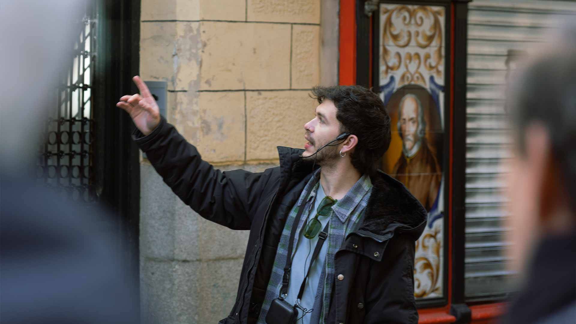 Un hombre señala algo mientras habla, de pie en una calle con edificios antiguos.