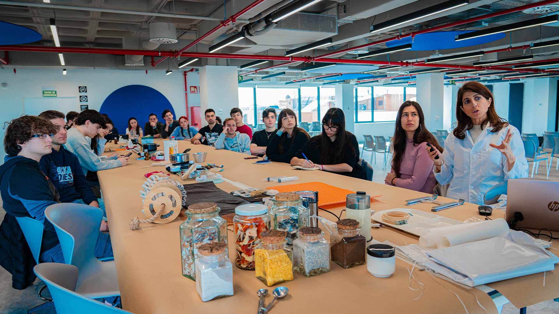 Un grupo de personas se reúne en una sala luminosa mientras una mujer habla y presenta ideas en una mesa llena de materiales diversos.