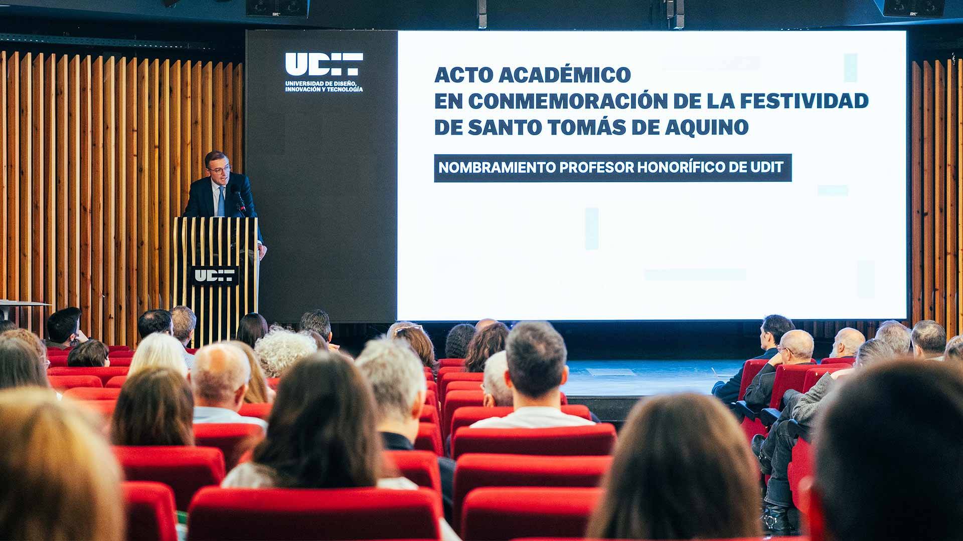 Un acto académico en conmemoración de la festividad de Santo Tomás de Aquino, con un orador frente a una audiencia en un auditorio moderno.