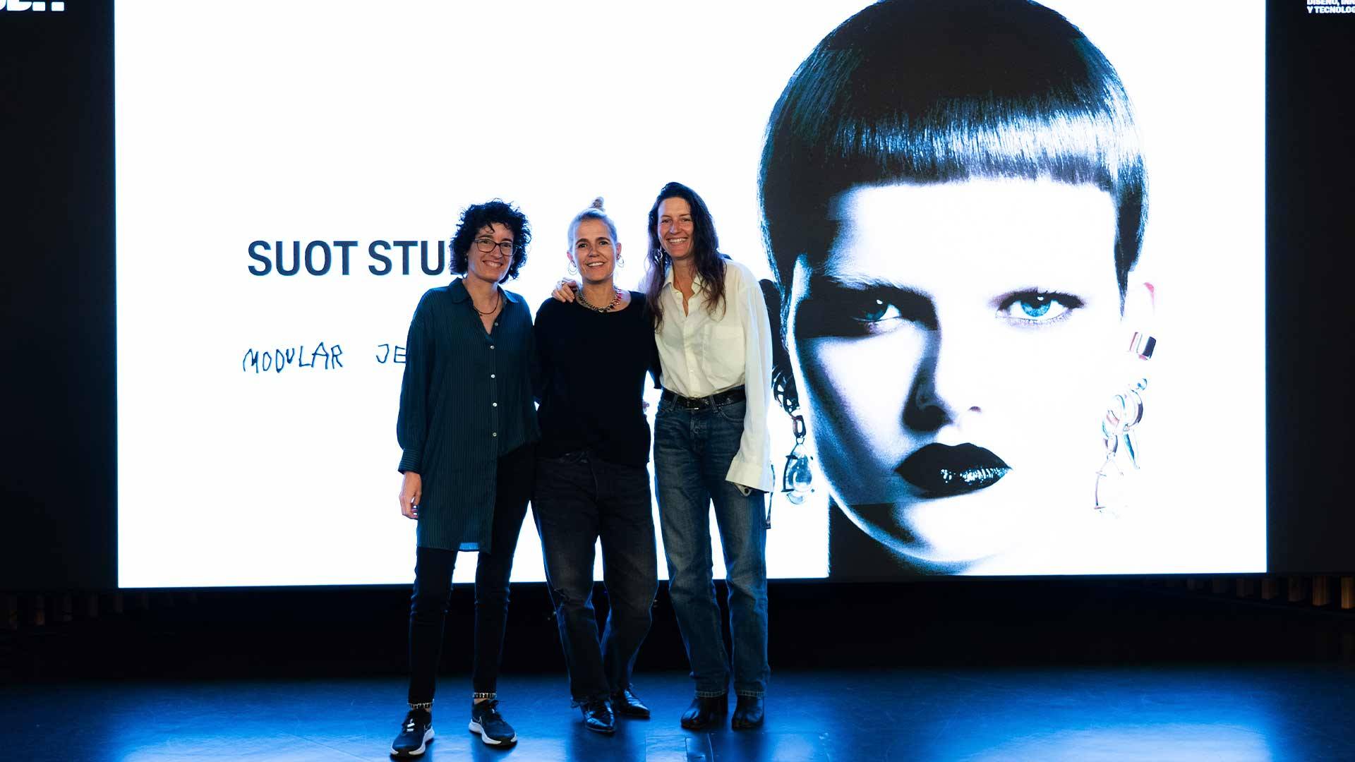 Tres mujeres posan frente a una pantalla grande con un rostro artístico y un logotipo.