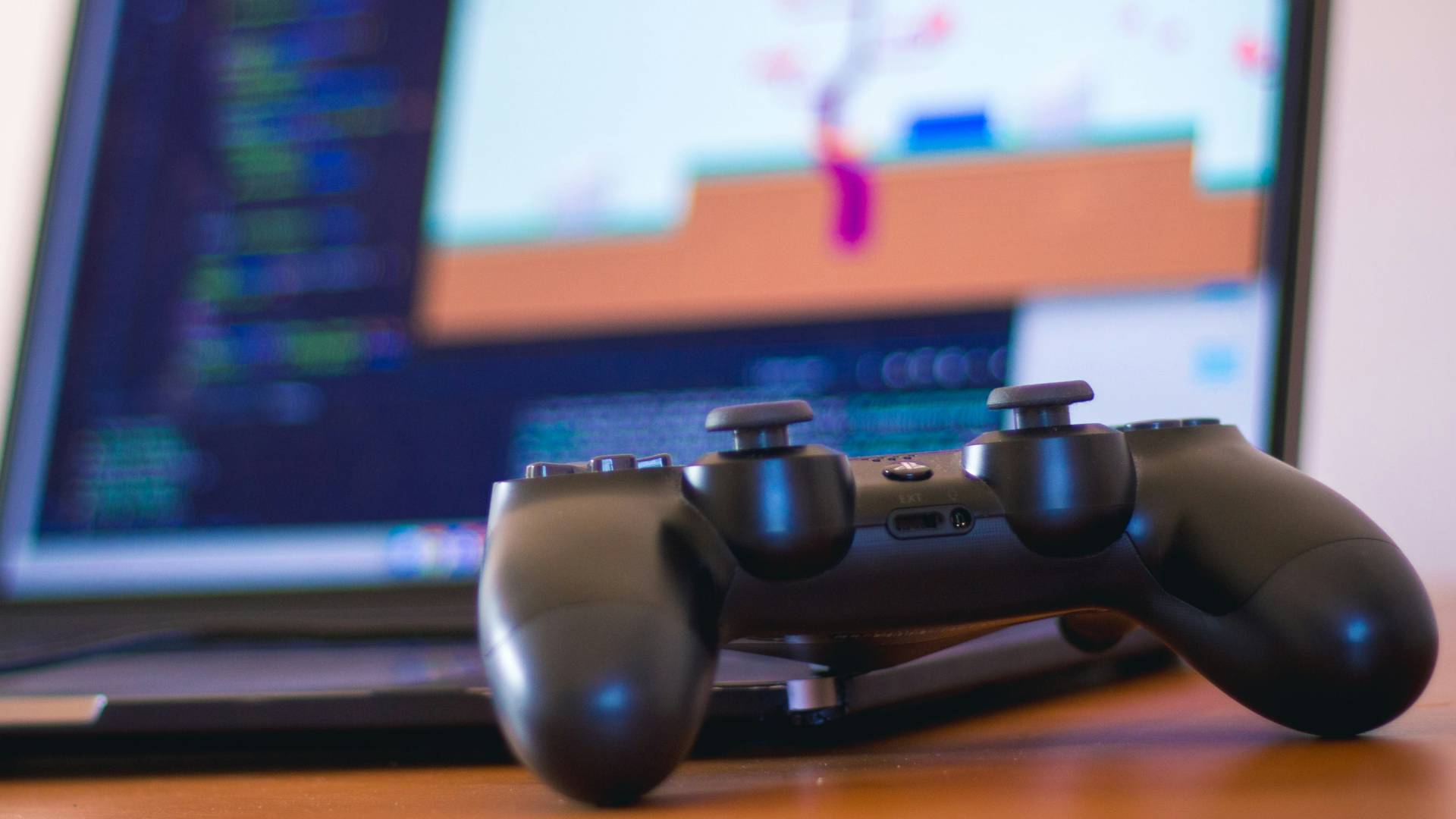 Una consola de videojuegos de tipo controlador se encuentra en primer plano frente a una computadora portátil que muestra una pantalla de programación.