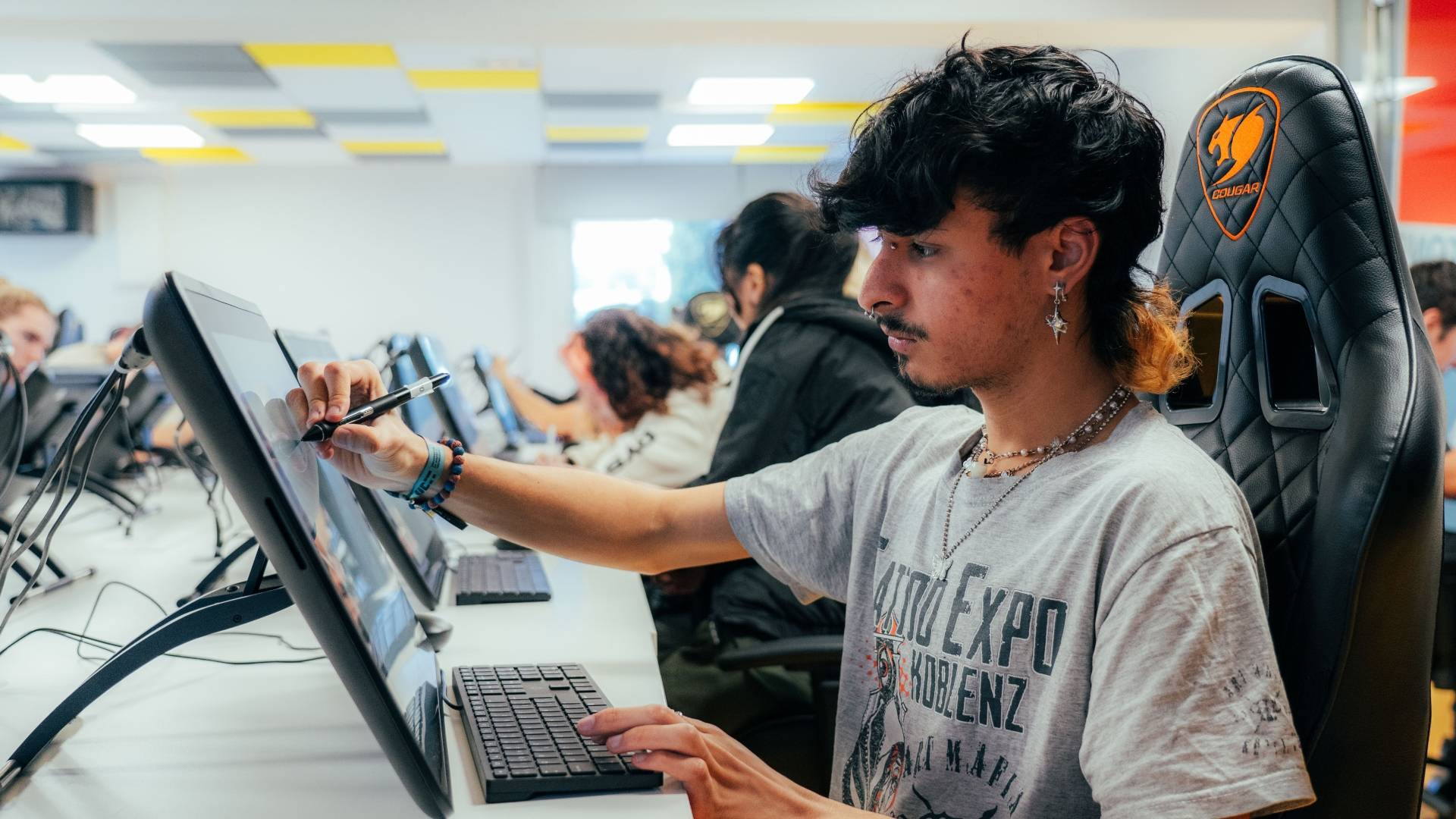 Un joven trabaja en una computadora digital en un ambiente de aula con otros estudiantes.