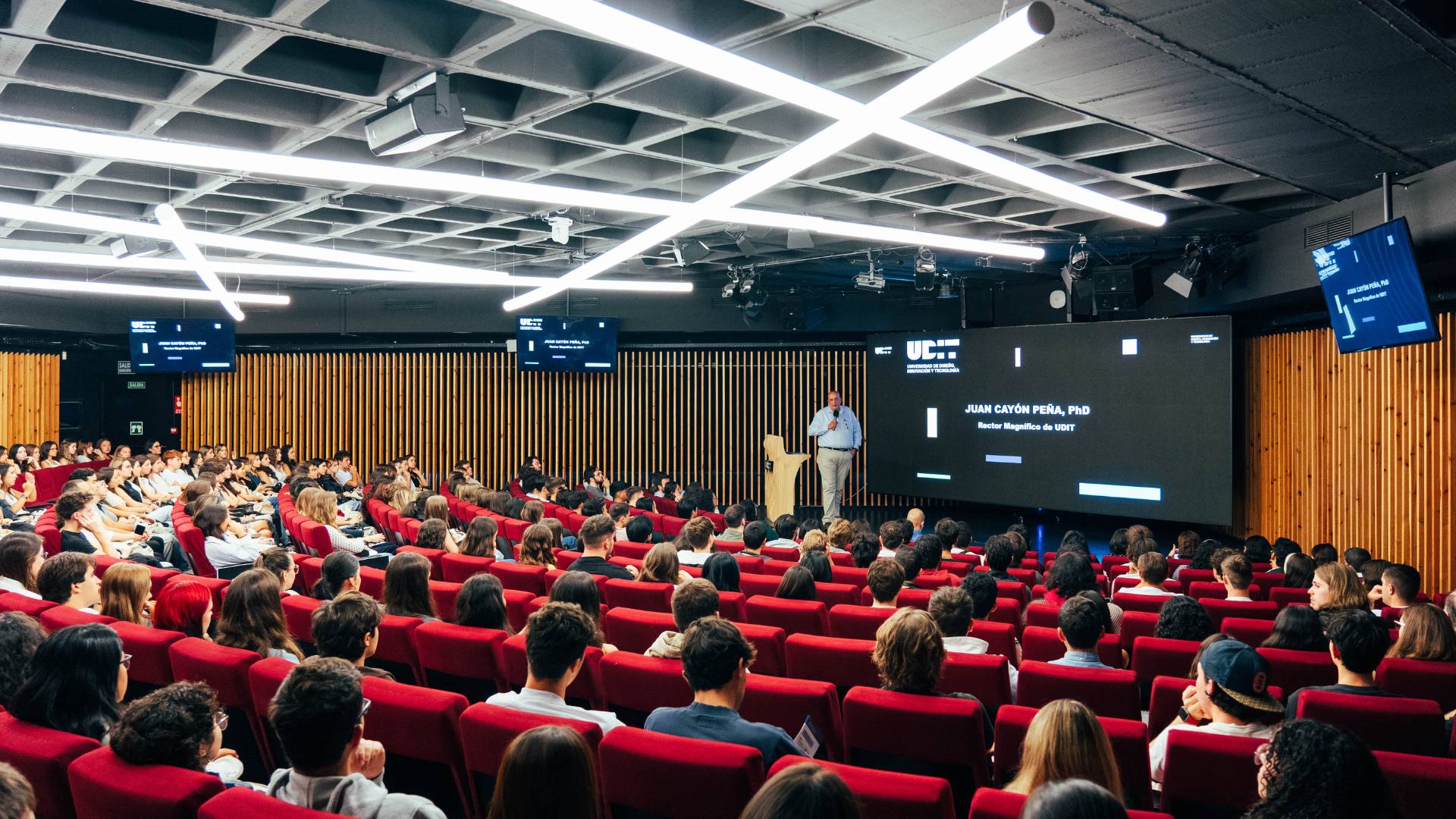 Un conferencista se presenta ante un gran auditorio lleno de estudiantes sentados en sillas rojas.