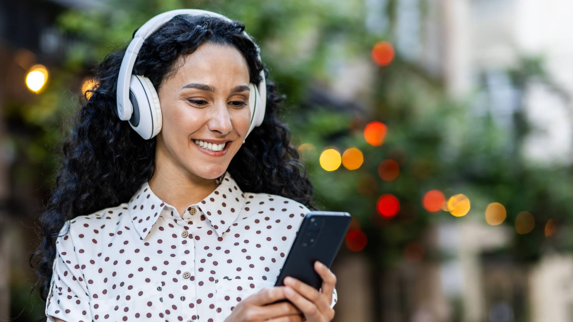 Una mujer sonriente con auriculares escucha música mientras usa su teléfono móvil en un entorno al aire libre.