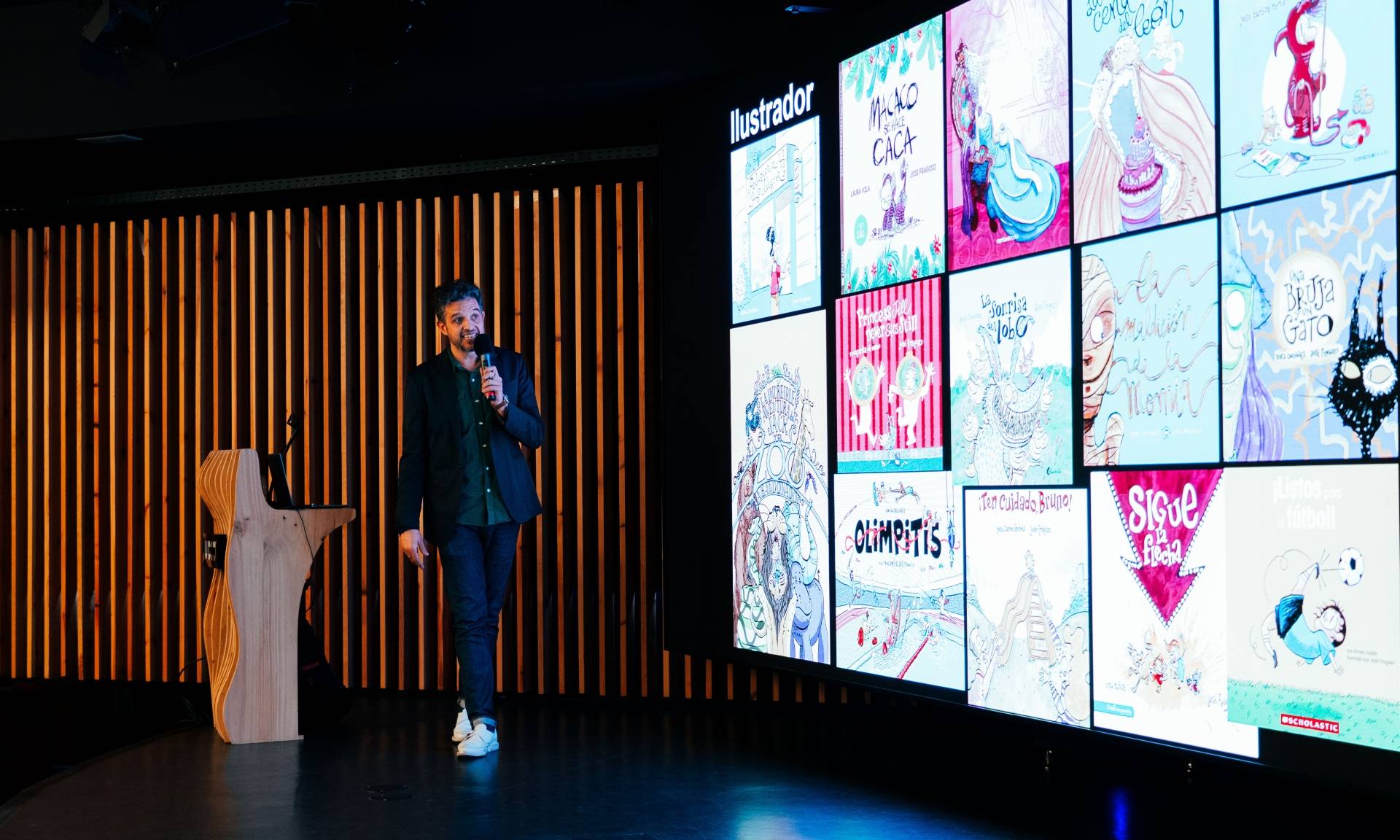 Un presentador habla frente a una pantalla que muestra diversas ilustraciones en un evento.