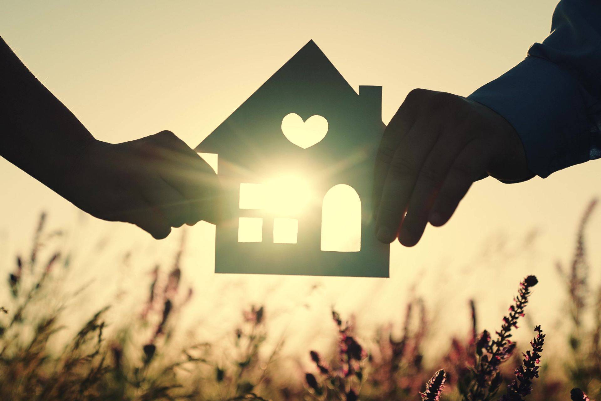 Dos manos sostienen una figura de una casa con un corazón mientras la luz del sol brilla a través de ella en un campo.