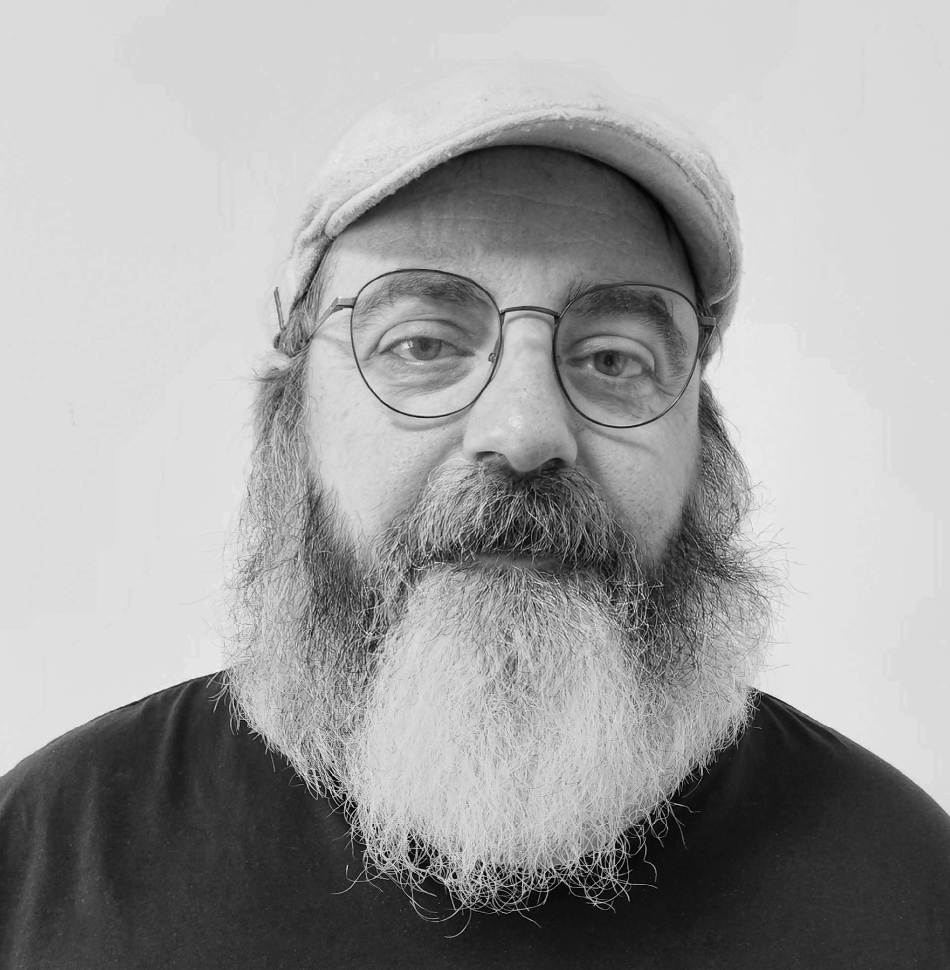 Un hombre con barba y gafas, vistiendo una camiseta negra y una gorra clara.