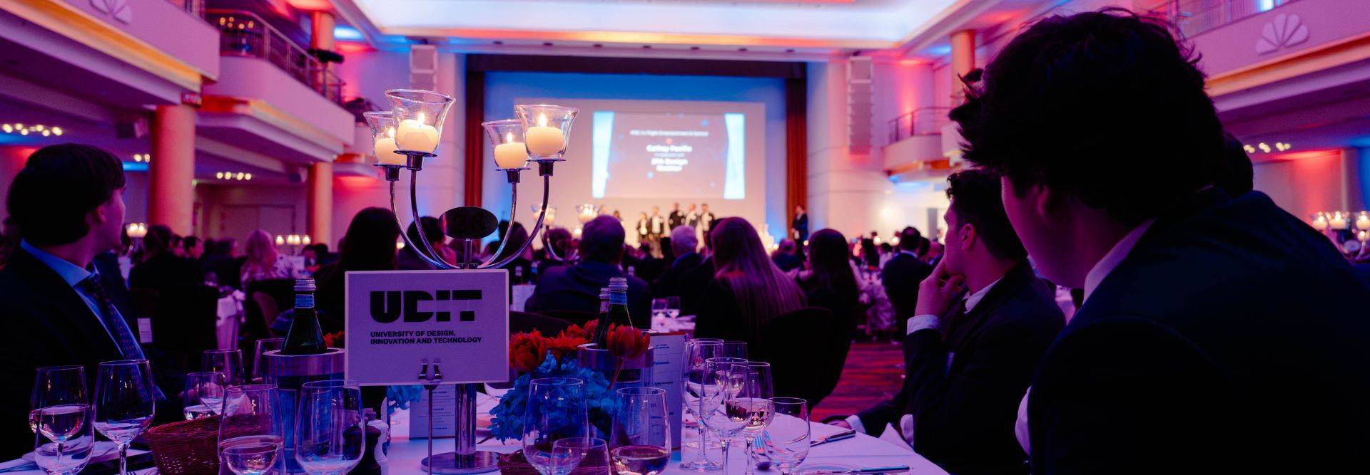 Una cena de gala con una presentación en un escenario iluminado y mesas decoradas.