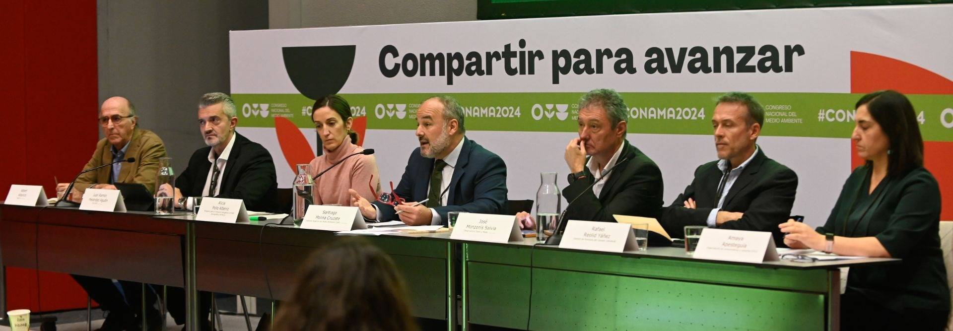 Una mesa redonda con siete personas sentadas frente a un panel que dice 'Compartir para avanzar'.