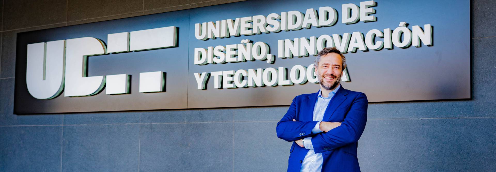 Un hombre sonriente posa frente a un letrero de una universidad que destaca la innovación y el diseño.