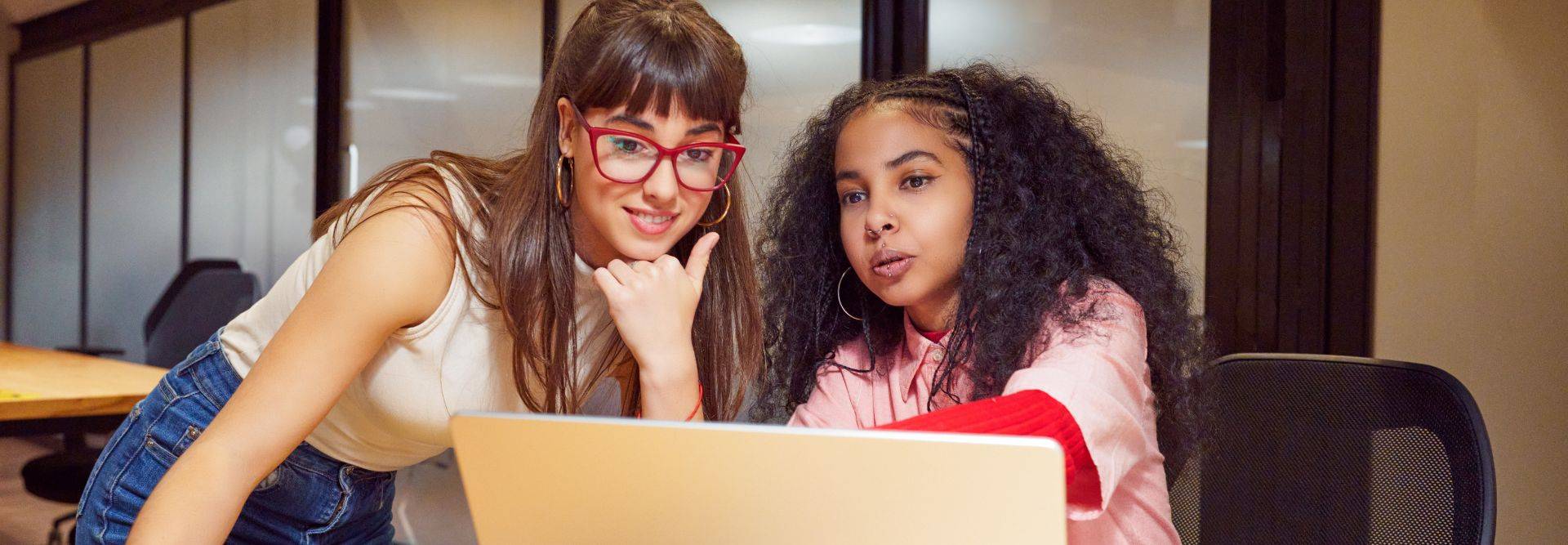 Dos mujeres están sentadas frente a una computadora portátil, revisando algo juntas con actitud concentrada.
