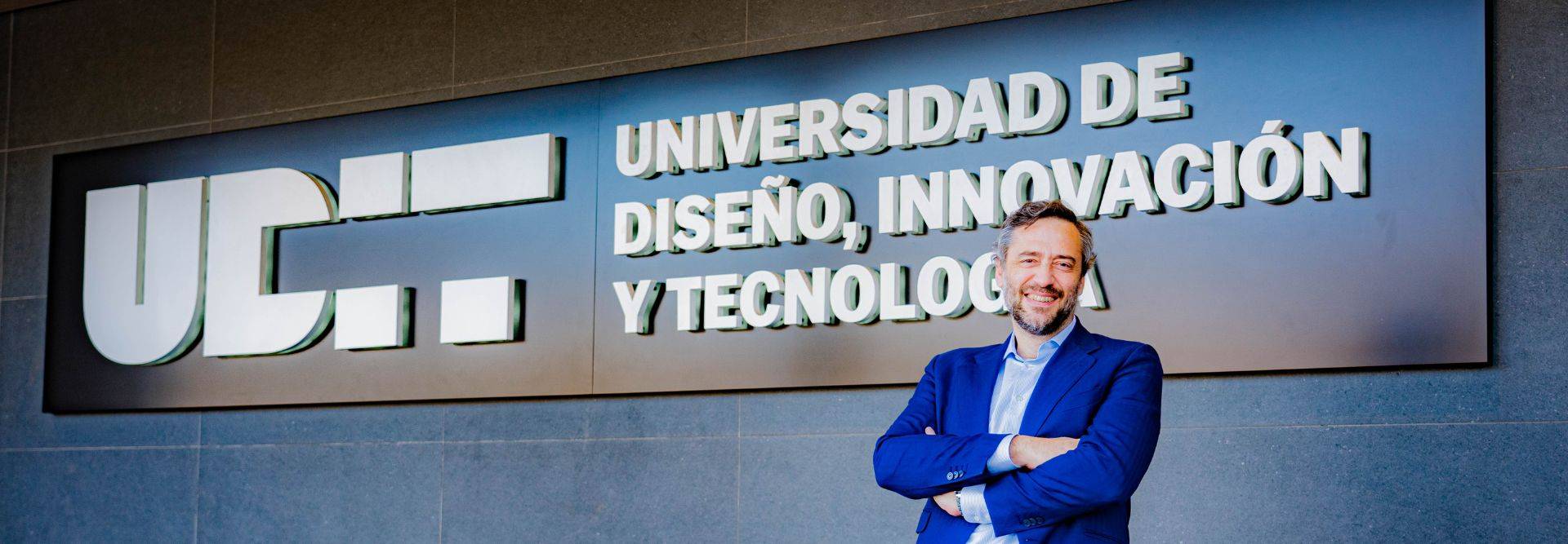 Un hombre sonriente se encuentra frente a un letrero de la Universidad de Diseño, Innovación y Tecnología.