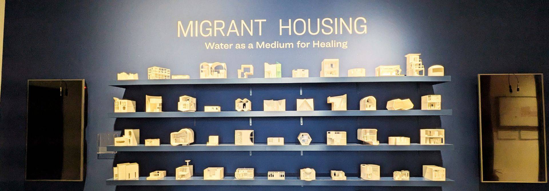 La imagen muestra una exhibición de maquetas sobre vivienda para migrantes en un fondo azul.