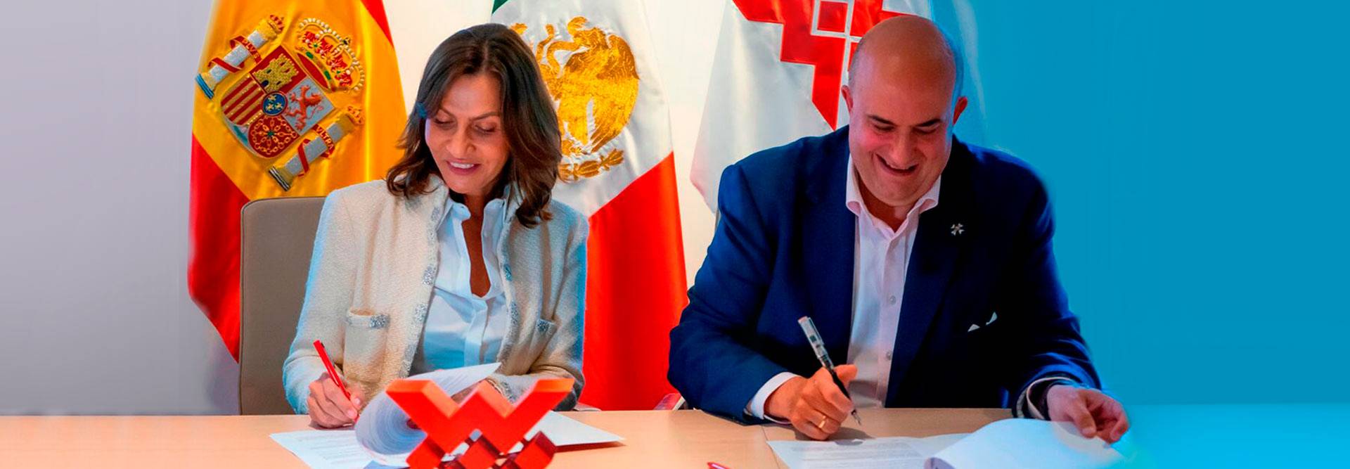 Dos personas firmando documentos en un evento oficial con banderas de España y México de fondo.