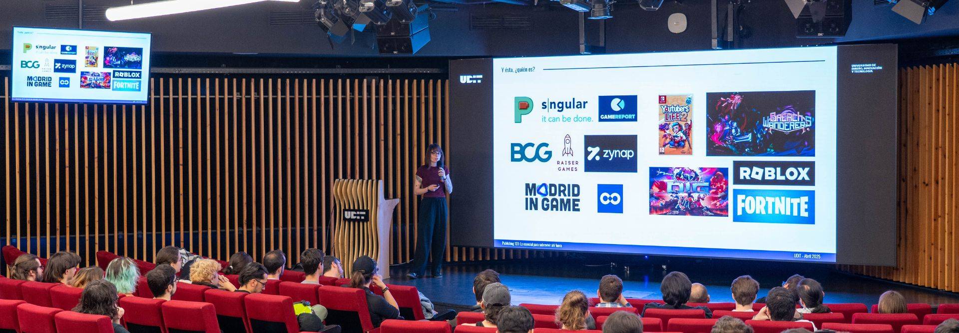Una presentación en un auditorio con un orador y un proyector mostrando logotipos de empresas relacionadas con tecnología y videojuegos.