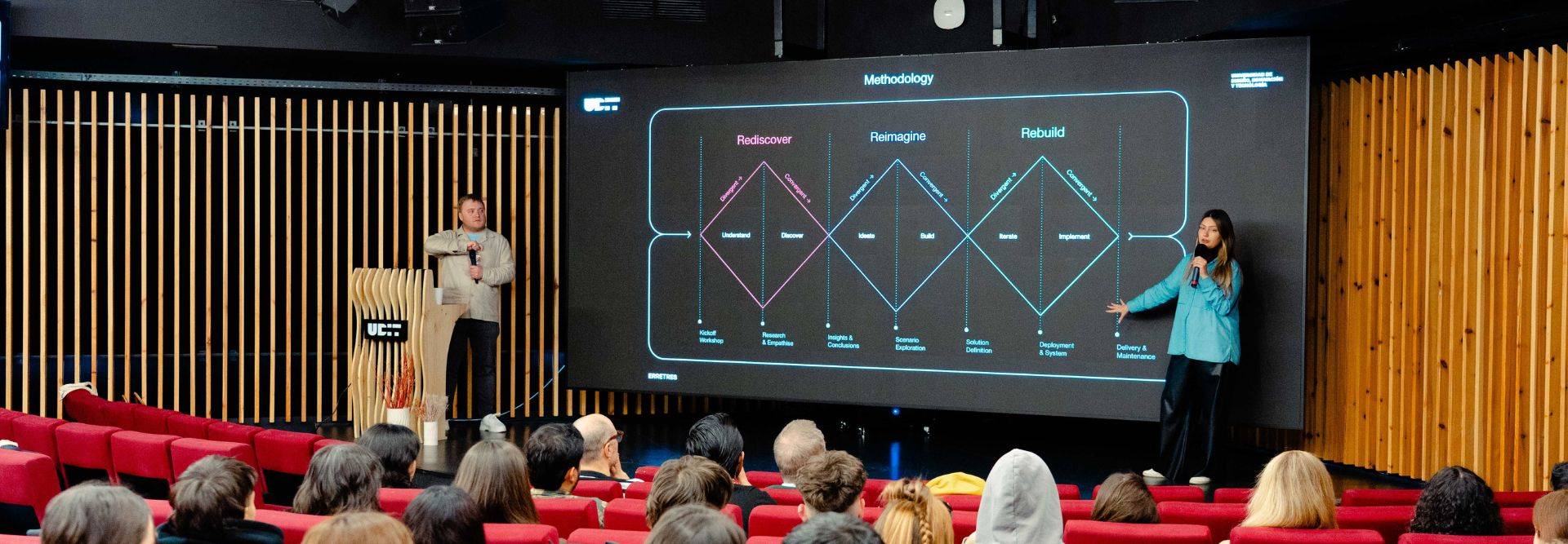 Una presentación en un auditorio con un gráfico en pantalla y dos ponentes interactuando con el público.