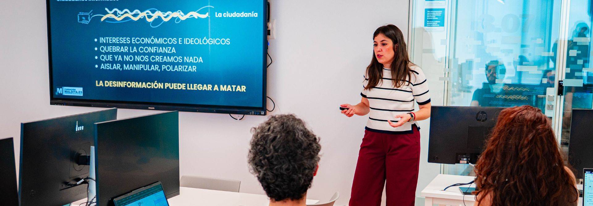 Una mujer expone sobre la desinformación en un aula con pantallas y alumnos.
