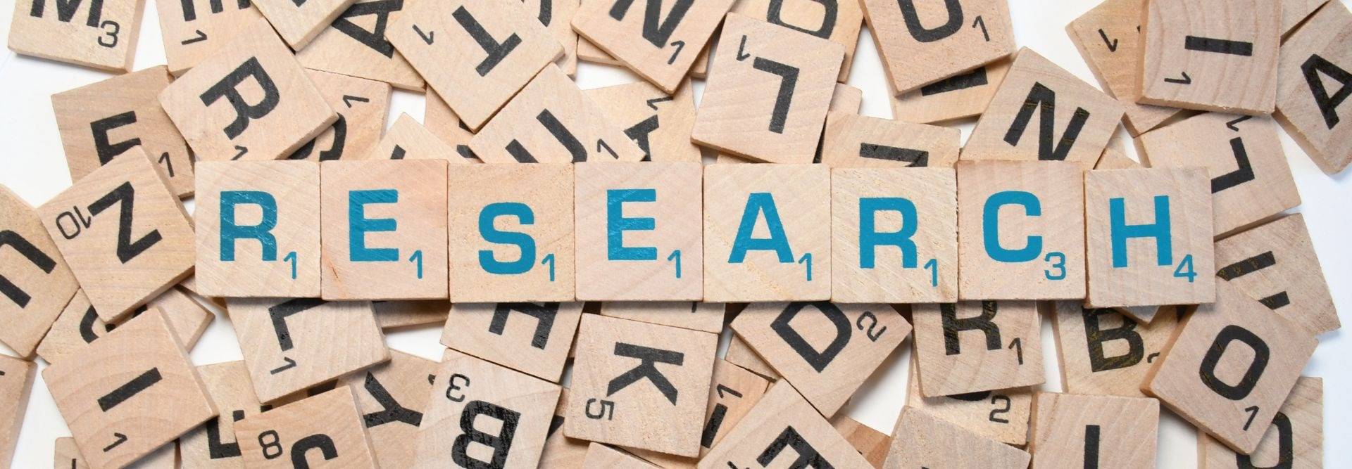 La imagen muestra letras de madera con la palabra 'RESEARCH' destacada en color azul entre letras de fondo.