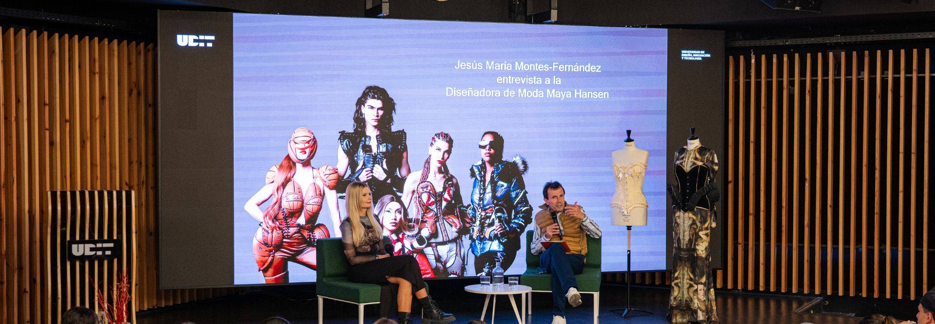 Imagen de un evento sobre diseño de moda con dos presentadores y un fondo artístico.