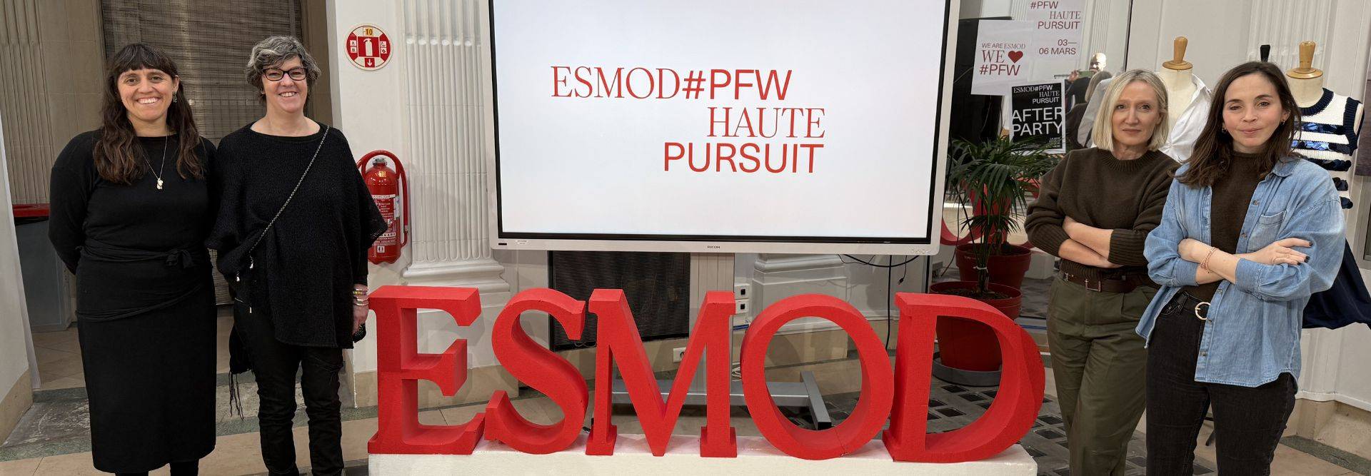 Cuatro mujeres se encuentran frente a un gran letrero que dice 'ESMOD#PFW HAUTE PURSUIT'.