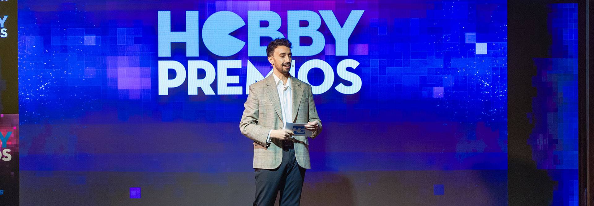 Un presentador en un evento de premios, de pie frente a un fondo decorado con el texto 'HOBBY PREMIOS'.