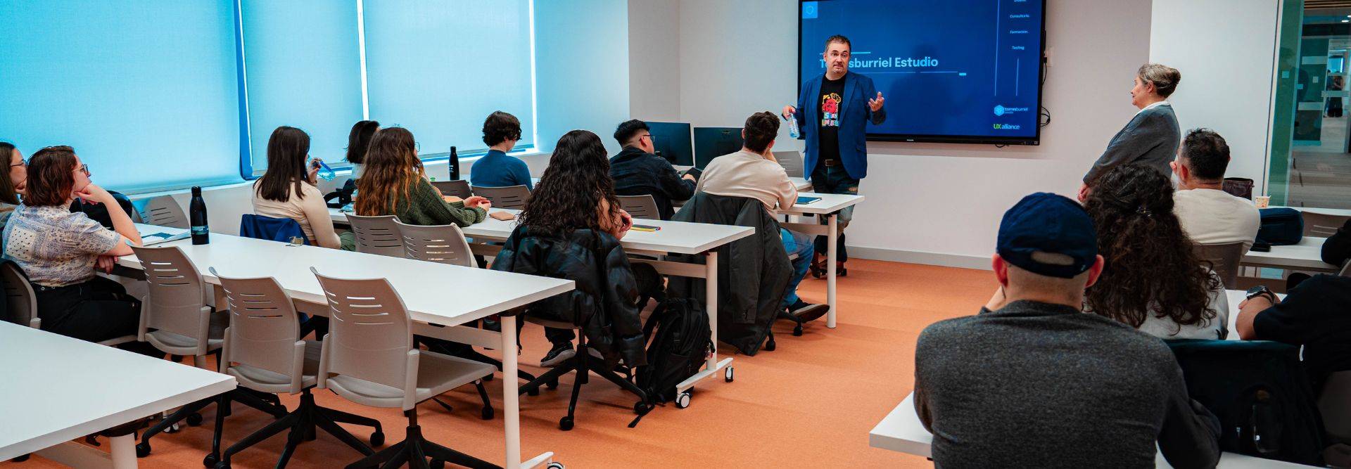 Una presentación en un aula con un orador frente a un grupo de estudiantes atentos.