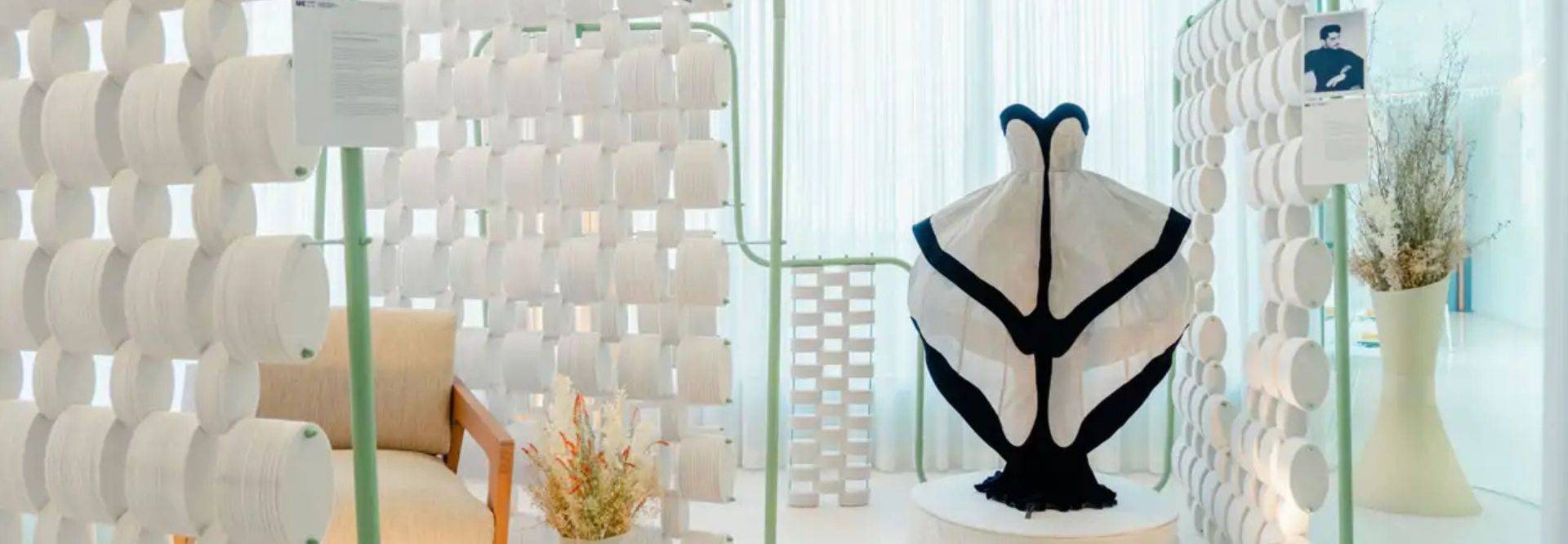 La imagen muestra un espacio interior con una escultura textil en el centro, rodeada de paneles decorativos y un sofá cercano.