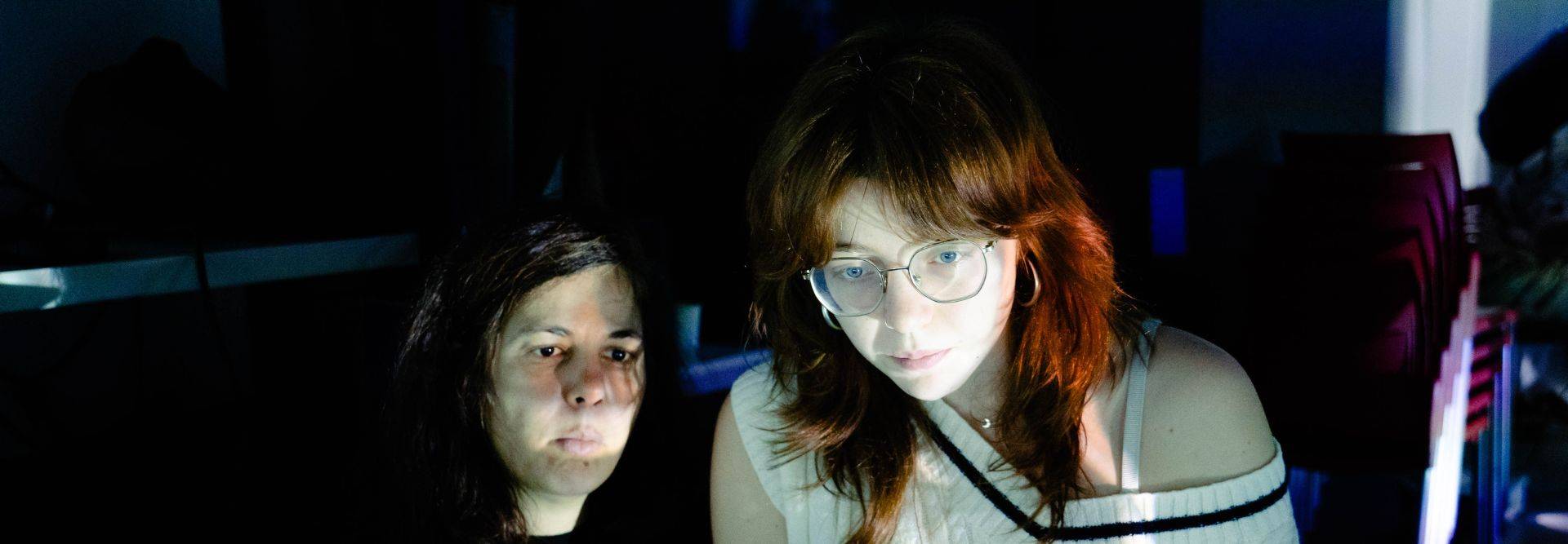 Dos personas están iluminadas por una luz tenue en un entorno oscuro.