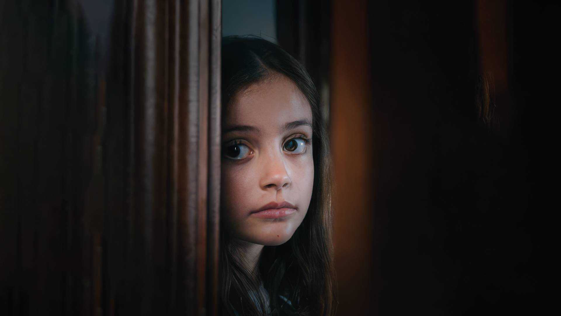 Una niña asoma su rostro tímidamente entre una puerta entreabierta.