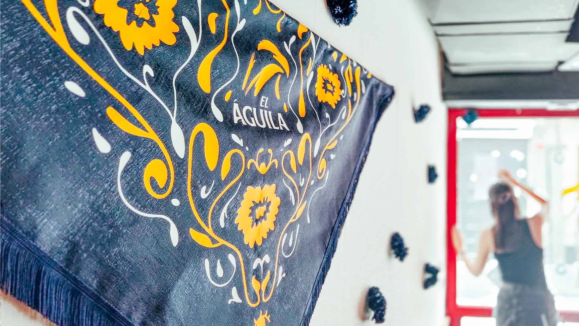 La imagen muestra un paño decorativo con flores amarillas y la palabra 'El Águila' en una pared blanca, mientras una persona trabaja al fondo.