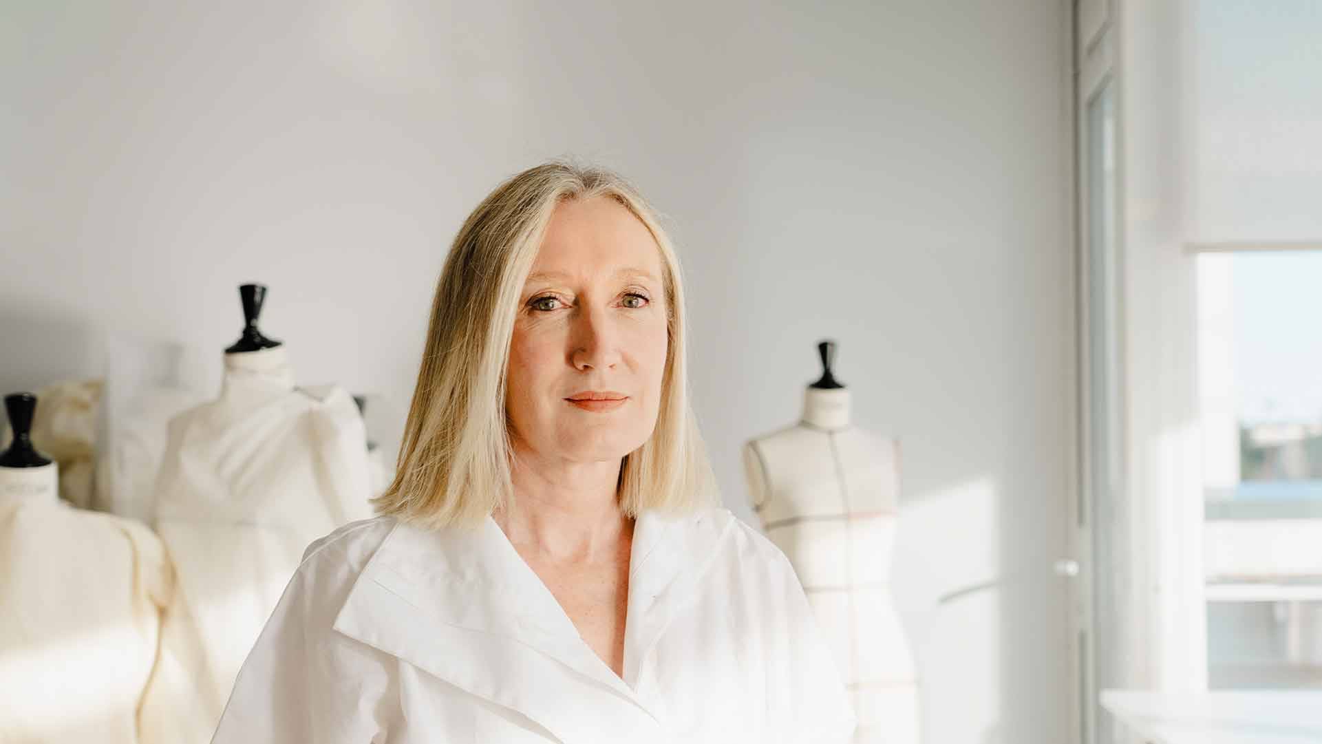 Una mujer de cabello largo y rubio, vestida con una blusa blanca, se encuentra frente a un fondo con maniquíes de moda.