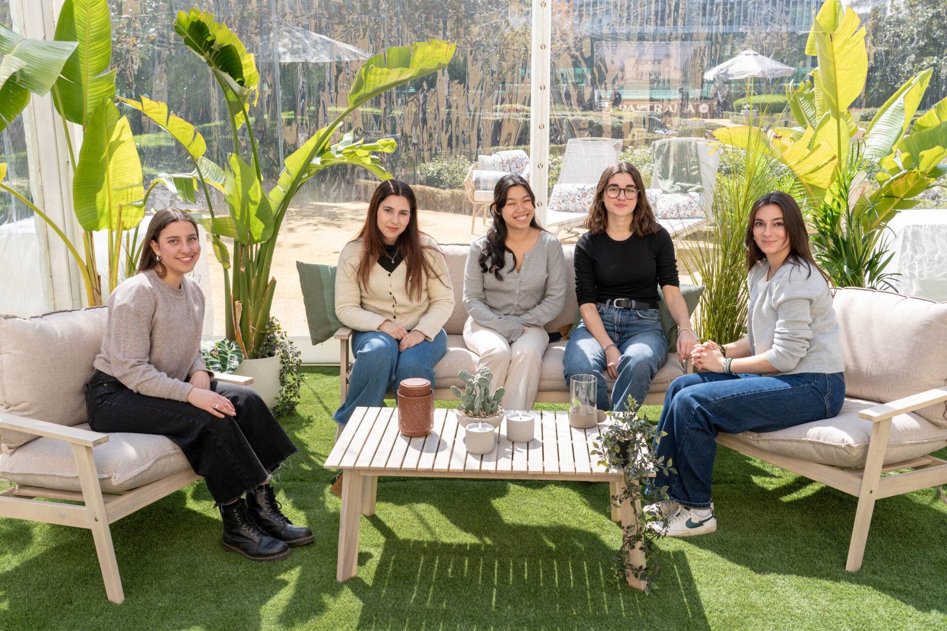 Cinco mujeres sentadas en un ambiente acogedor y soleado, rodeadas de plantas.