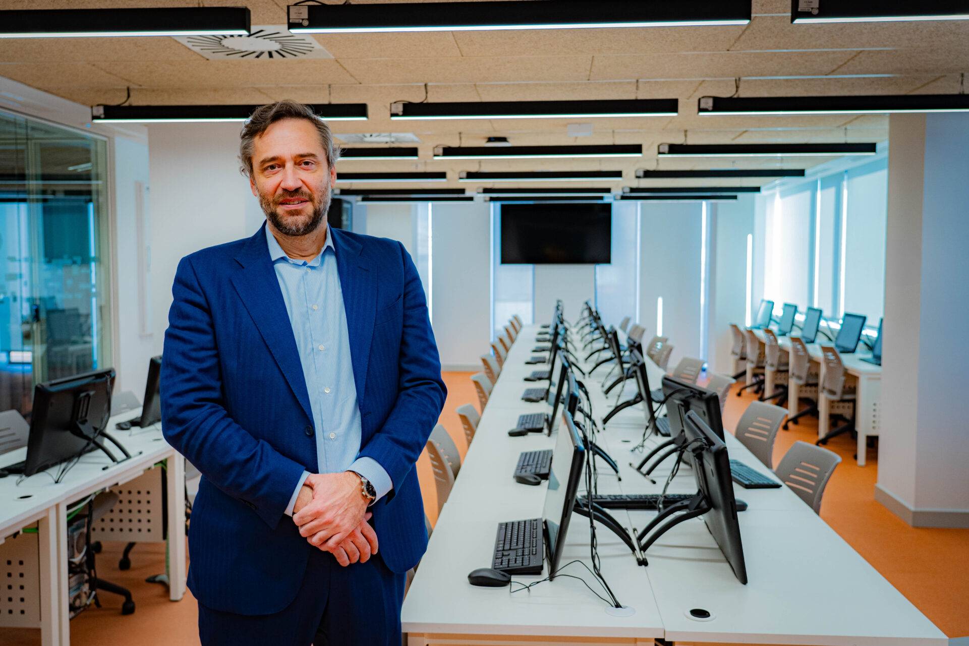 Un hombre de pie en un aula moderna con mesas y computadoras desocupadas.