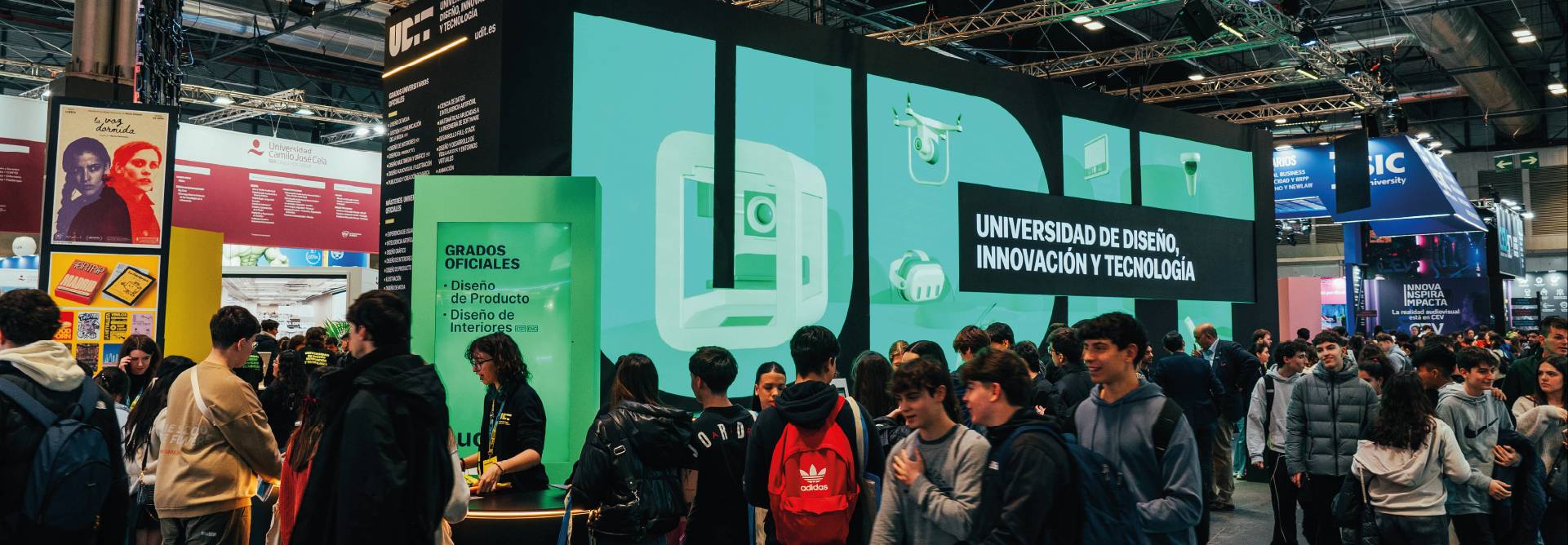 Una multitud de personas en una feria educativa frente a un gran stand que representa una universidad de diseño e innovación.