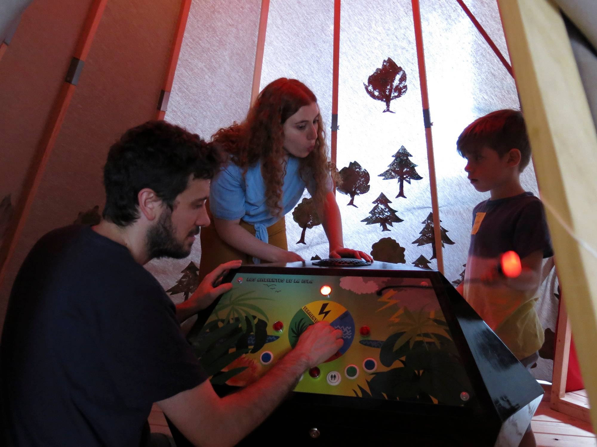 Un hombre, una mujer y un niño juegan juntos en un entorno recreativo dentro de una cabaña decorada con árboles recortados.