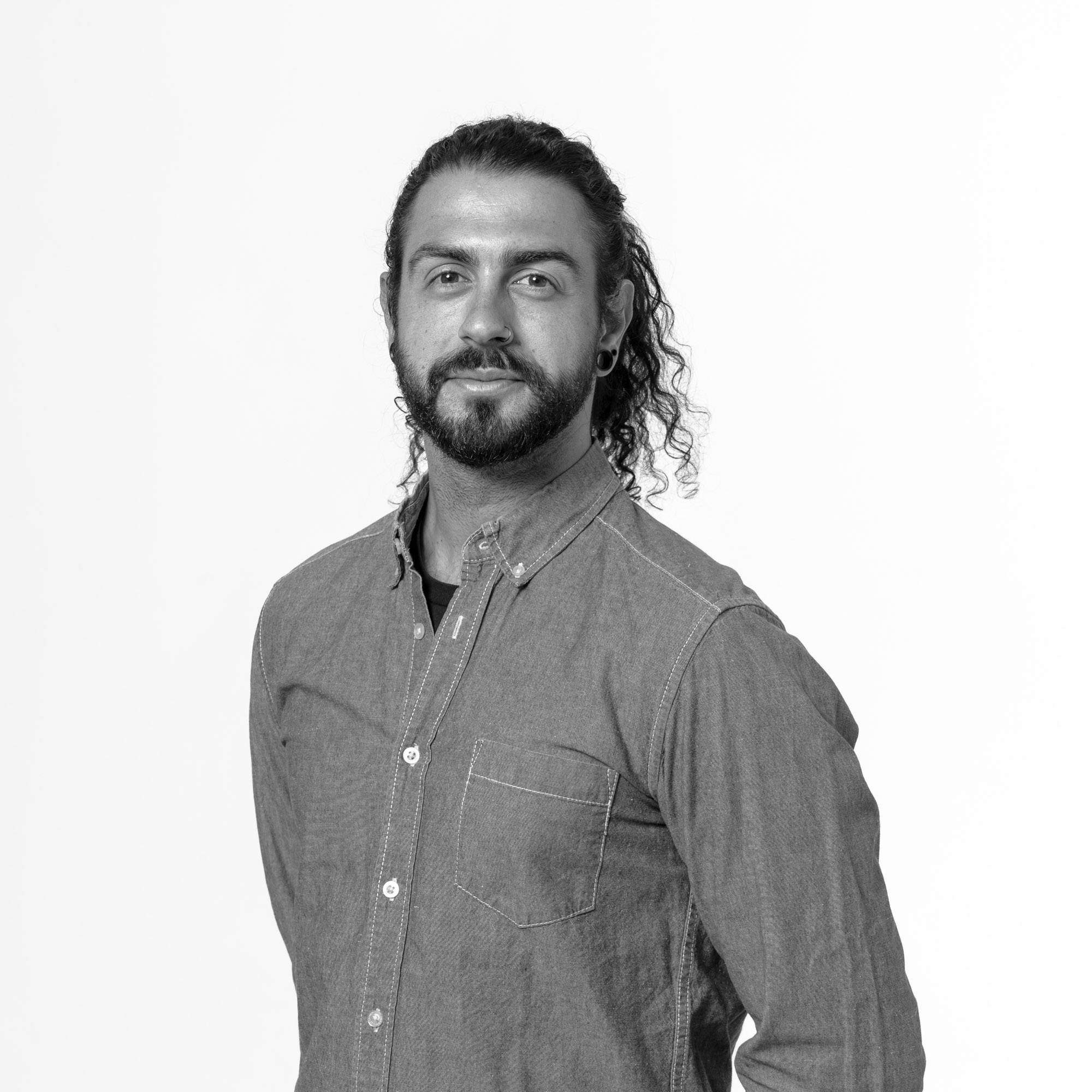 Un hombre con barba y cabello largo, vestido con una camisa de mezclilla, posa frente a un fondo blanco.