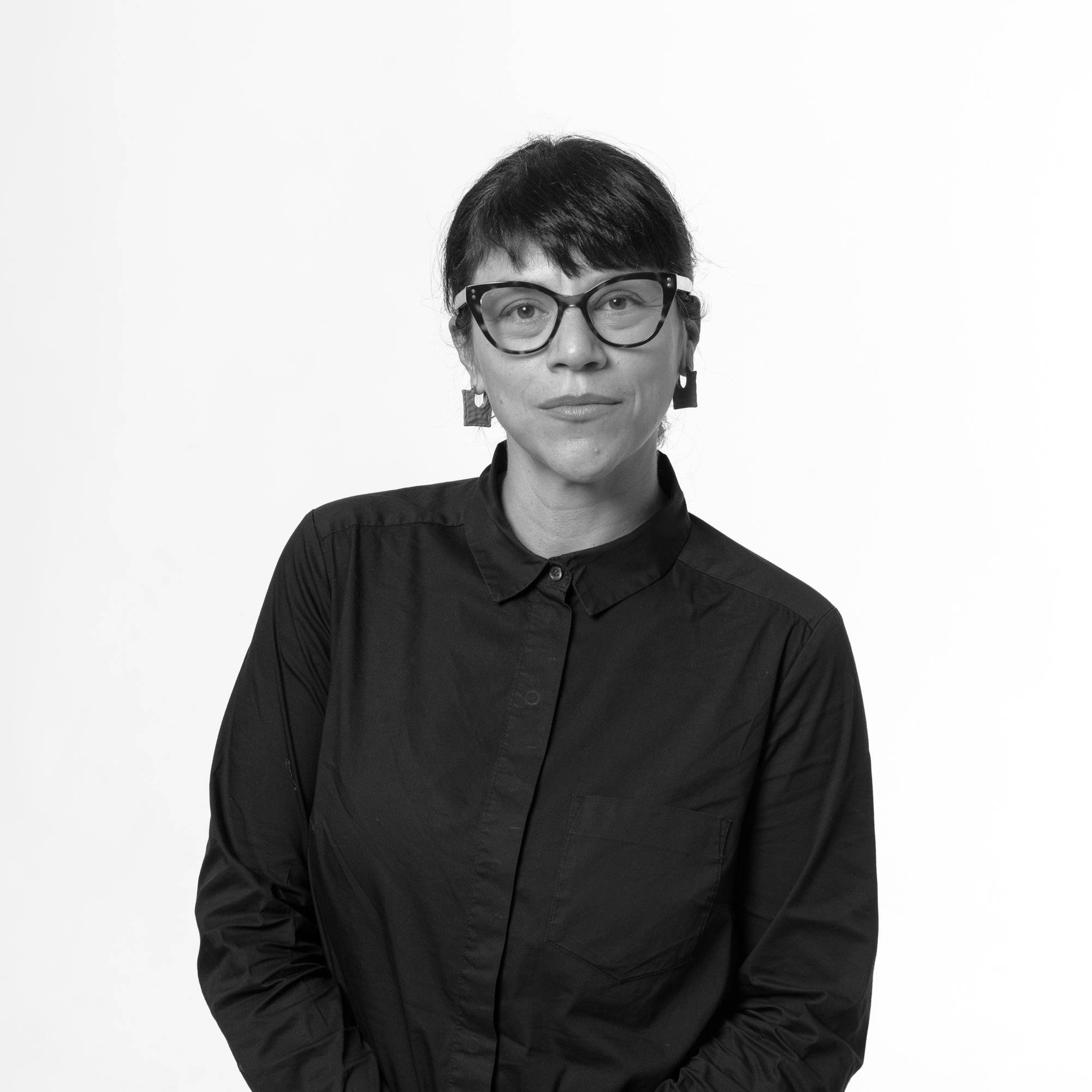 Mujer con gafas y blusa negra posa en un fondo blanco.